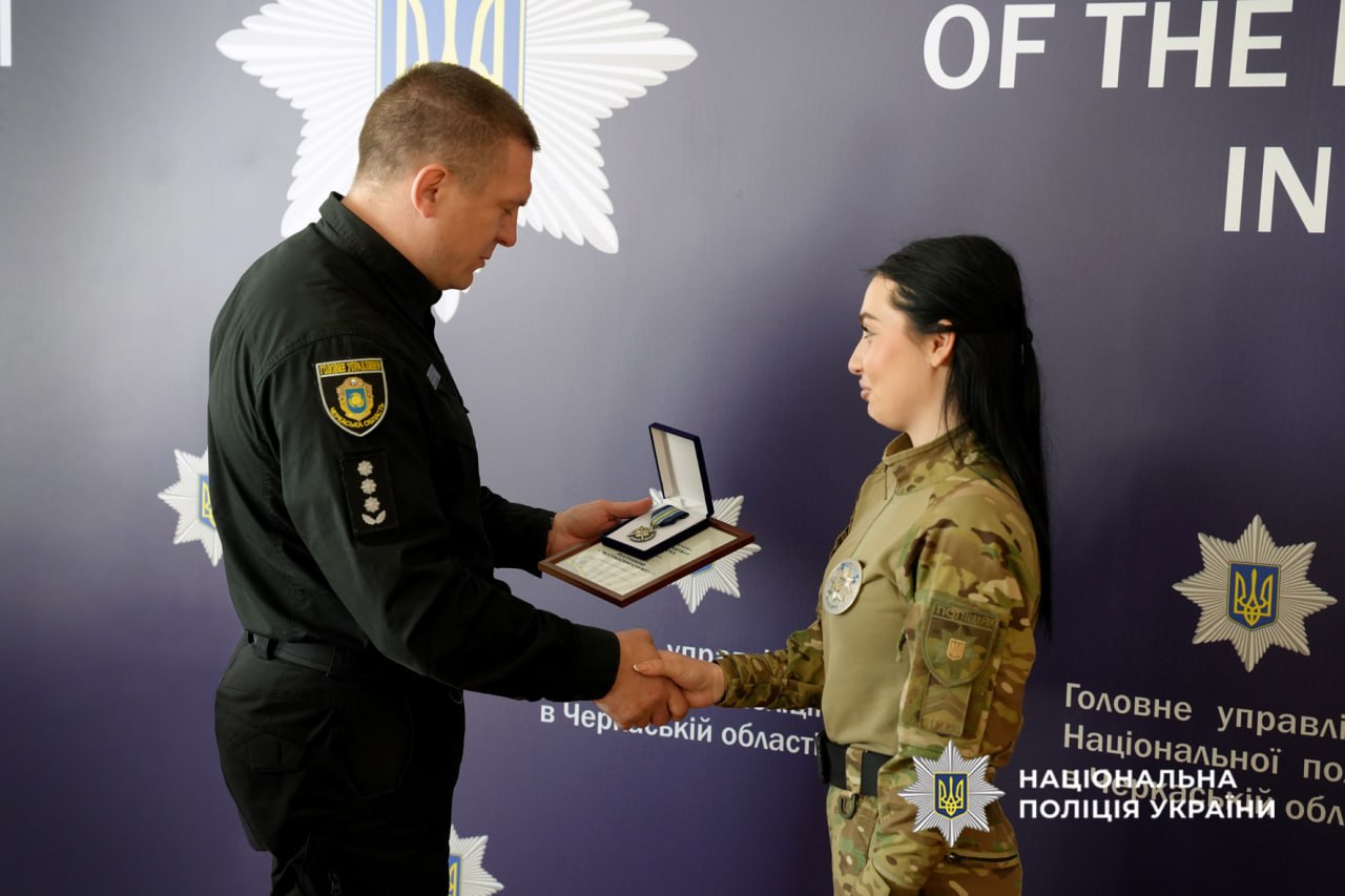 Male officer in black uniform presents a medal to a female soldier in camouflage while they shake hands in a police backdrop.