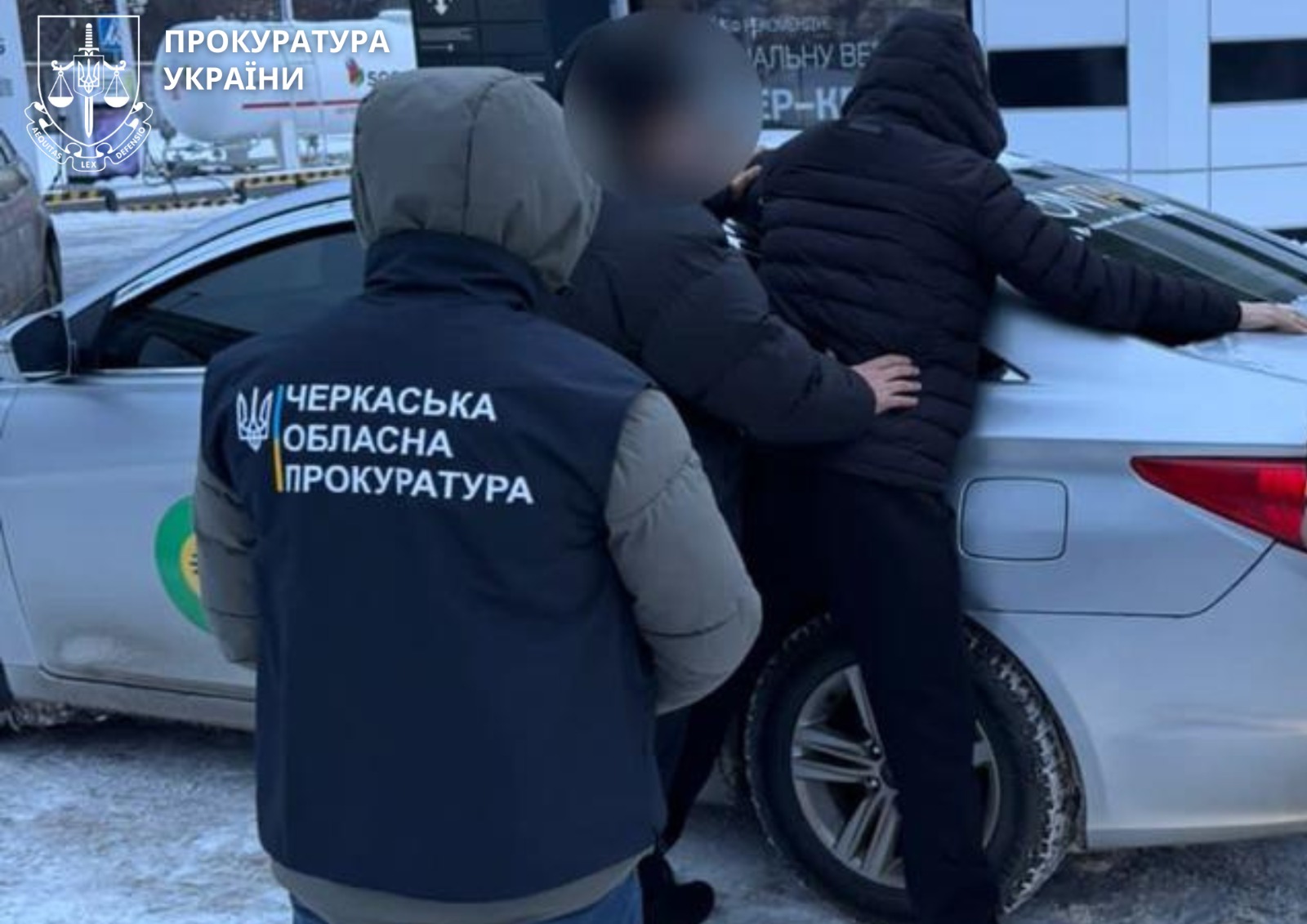 Officers from the Cherkasy Regional Prosecutor's Office detain a person next to a silver car in a snowy area; one officer has a jacket with Ukrainian emblem and text on the back.