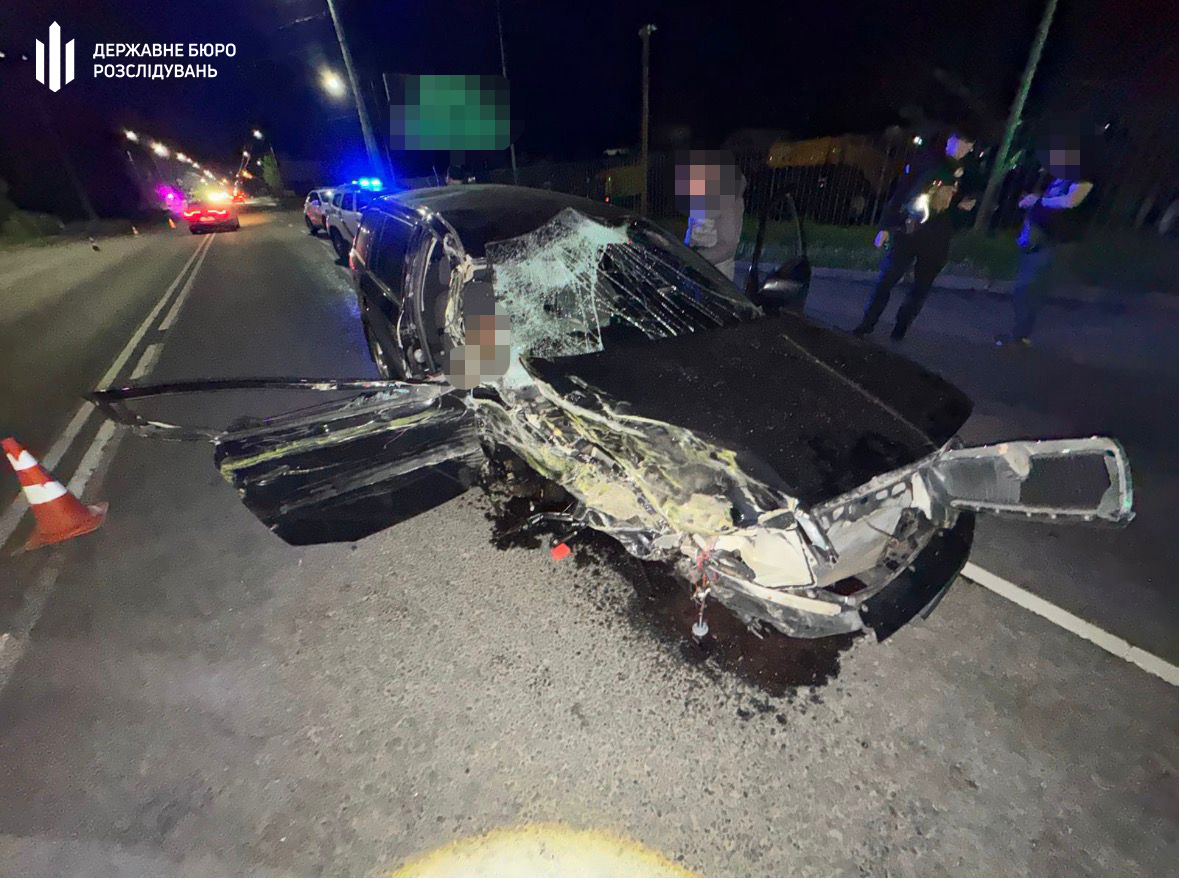 Nighttime car crash scene: a black sedan with extensive front-end damage and a shattered windshield, police car with flashing blue lights, and bystanders on a dimly lit street.