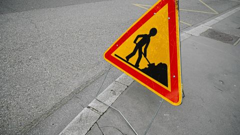 Triangular yellow road work sign with a worker icon placed on a sidewalk near a curb, warning of construction ahead.