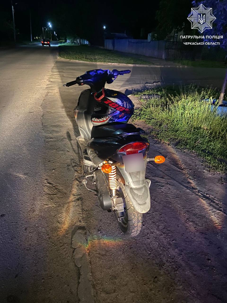Motorcycle parked on a dimly lit street at night, helmet with a shark-mouth graphic on the seat, police logo in the top-right corner indicating Ukrainian Patrol Police.
