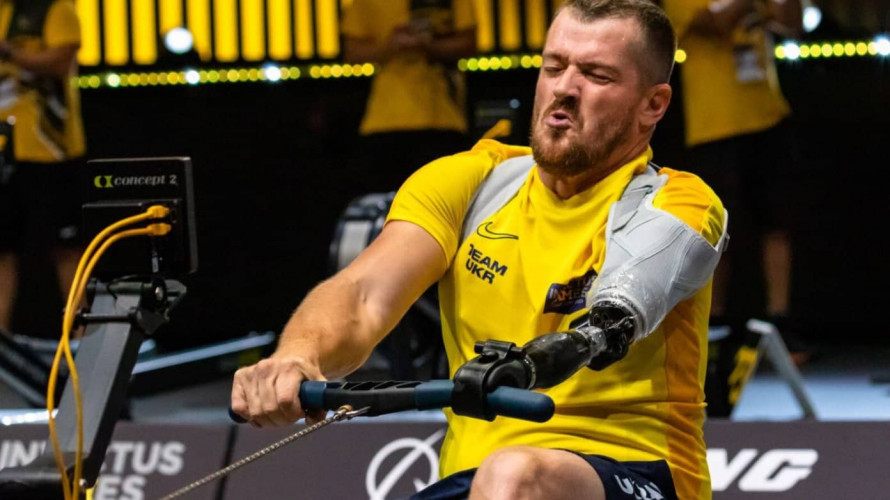 Male para-athlete in a bright yellow shirt pulls on a resistance bar, using a prosthetic left arm during a competition, showing intense effort.