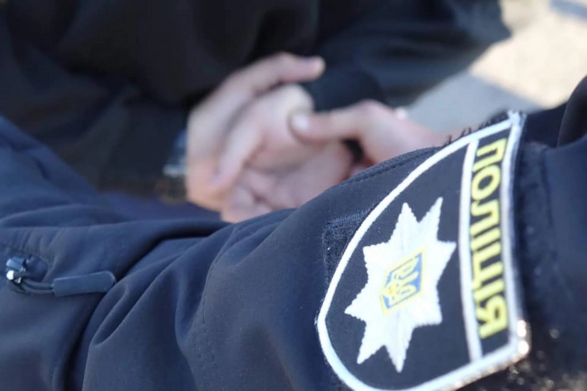 Close-up of a dark uniform sleeve with a shield-shaped insignia patch, featuring a white border and yellow lettering, partially visible on the right side.