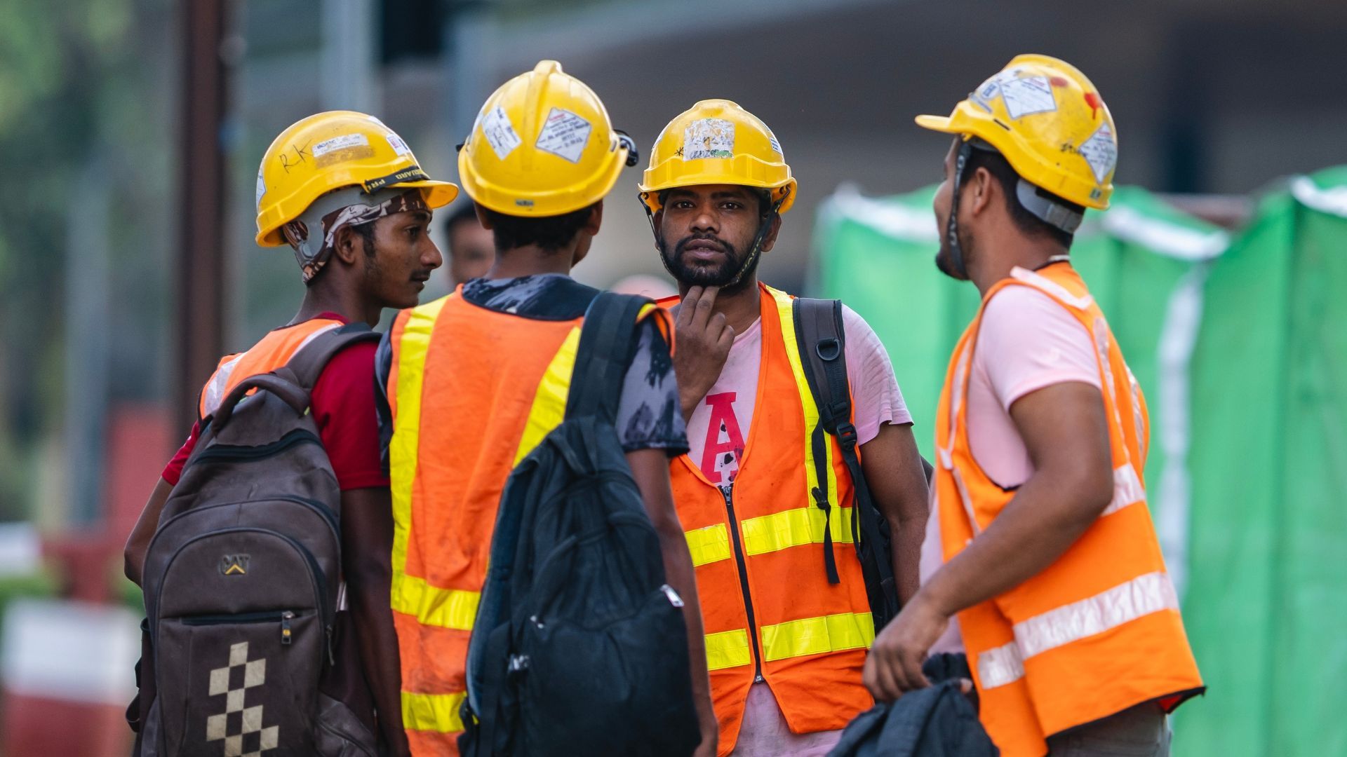 Construction workers in yellow hard hats and orange reflective vests talking on a site.