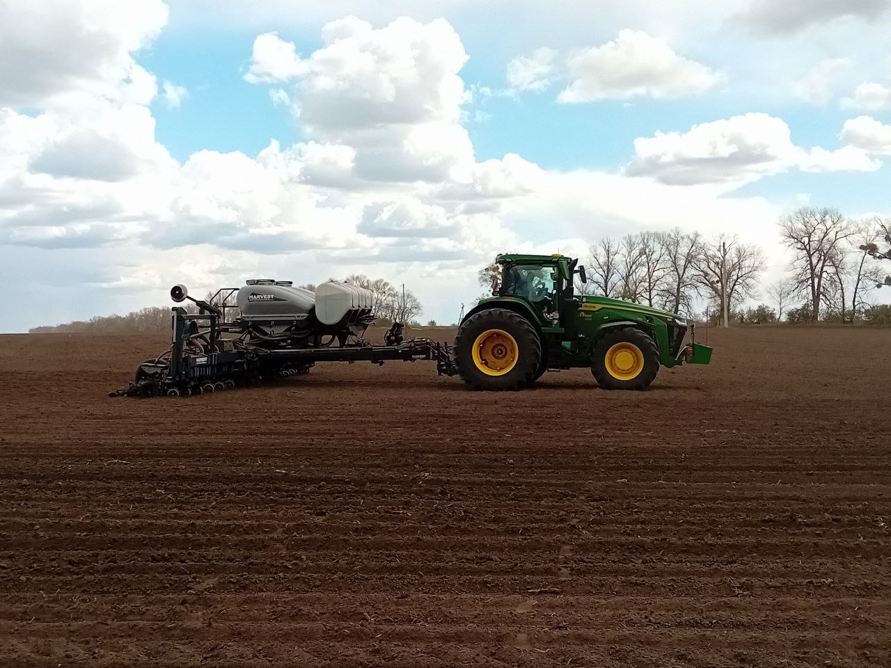 Green tractor pulling a large seed planter across a plowed field under a blue sky with clouds