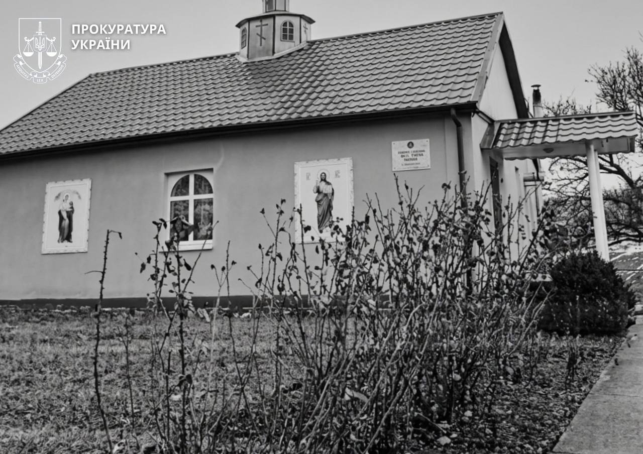 Black-and-white photo of a small church-like building with tiled roof and religious icons on its exterior walls, foreground shrubs, and a Ukraine Prosecution logo in the top-left corner.