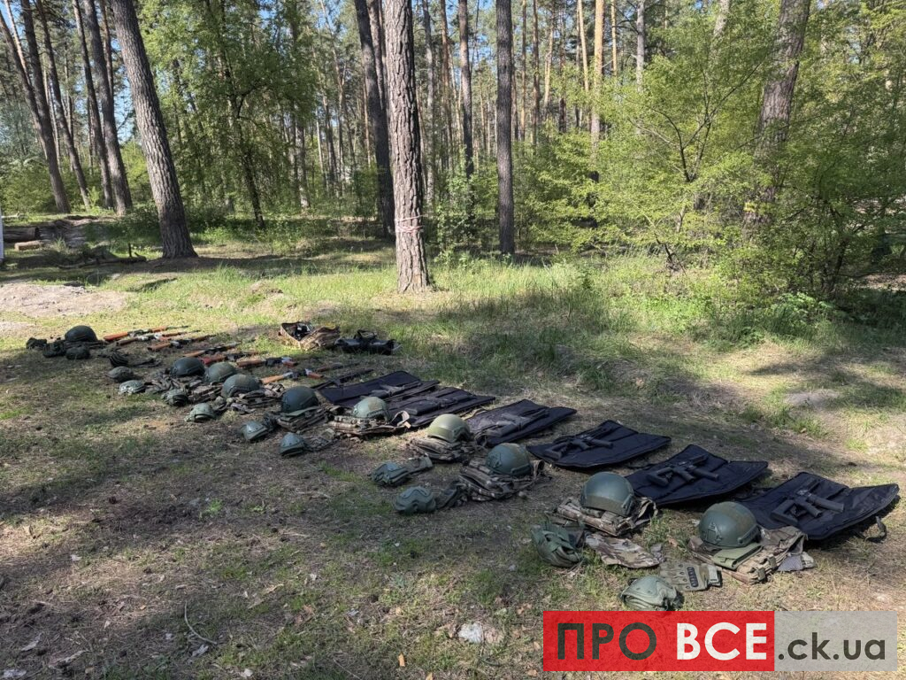 Line of green military helmets, vests, and packs laid out on the grassy forest floor beneath tall trees, organized in rows.