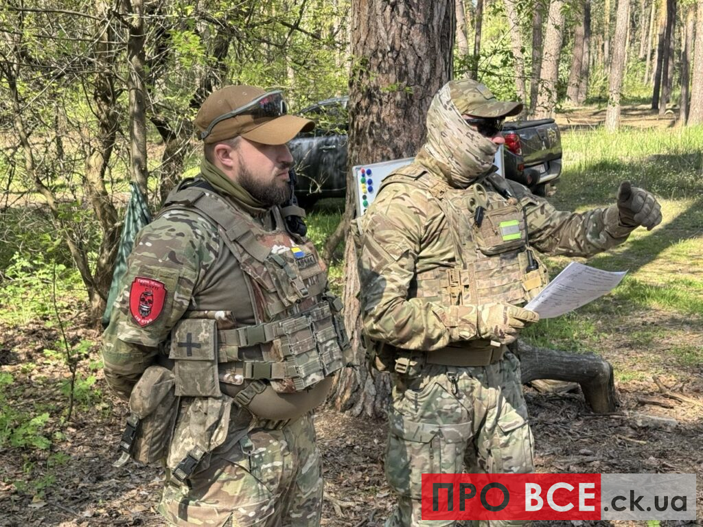Two soldiers in woodland camouflage gear stand in a forest; one has a red shield patch on his sleeve.