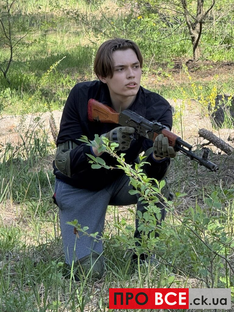 Young person kneeling in a grassy outdoor area, holding a rifle with a wooden stock, looking to the side.