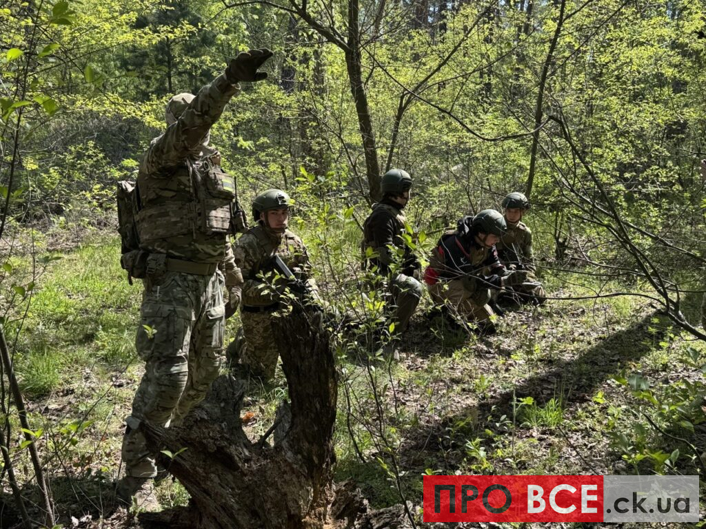 Soldiers in camouflage gear in a forest, one man standing and signaling with his arm while others crouch and observe a training exercise.