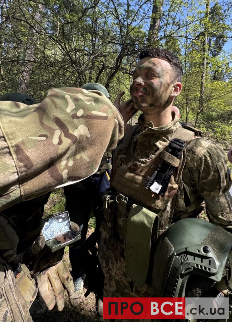 Soldier in camouflage being painted with green face paint by a comrade during an outdoor training exercise in a forest.