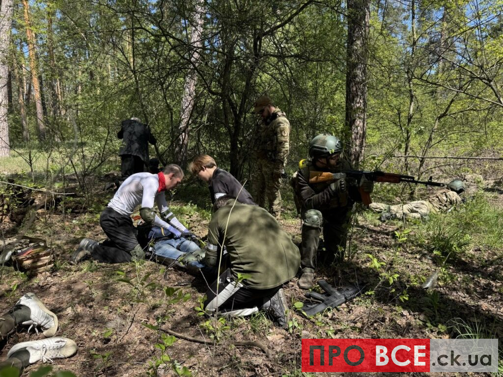 Medics treat a wounded person on the forest floor while soldiers stand and kneel nearby during a training exercise in the woods.