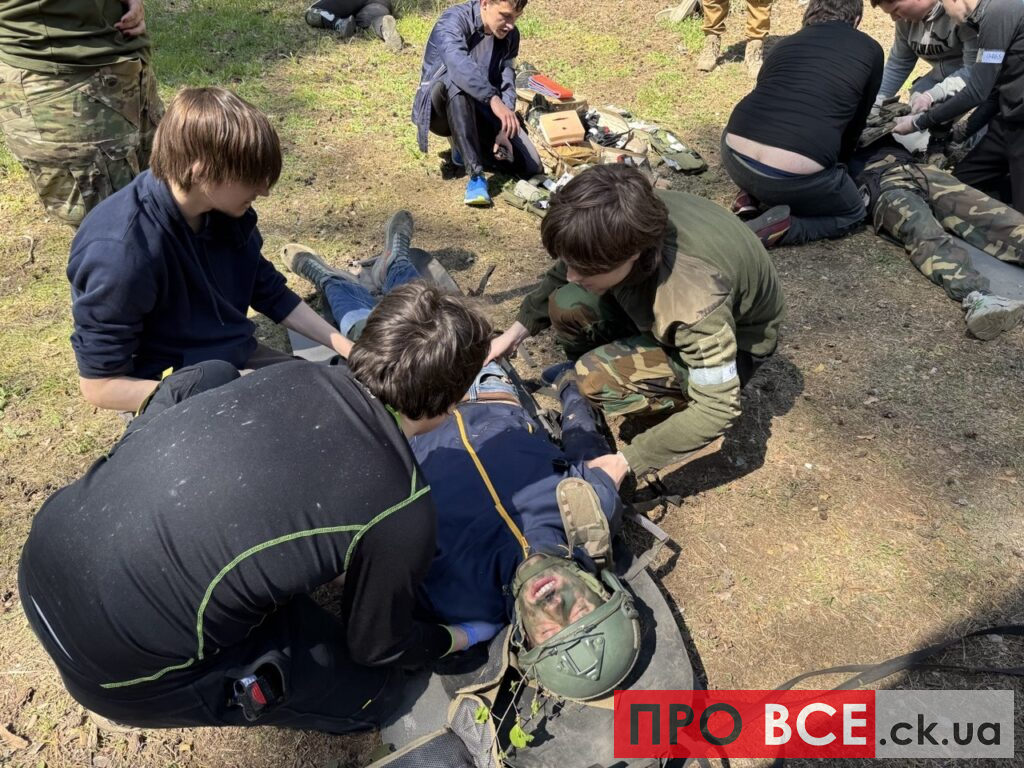 Group of trainees in outdoor gear attend to a man lying on the ground with a helmet, practicing a medical rescue drill outside.