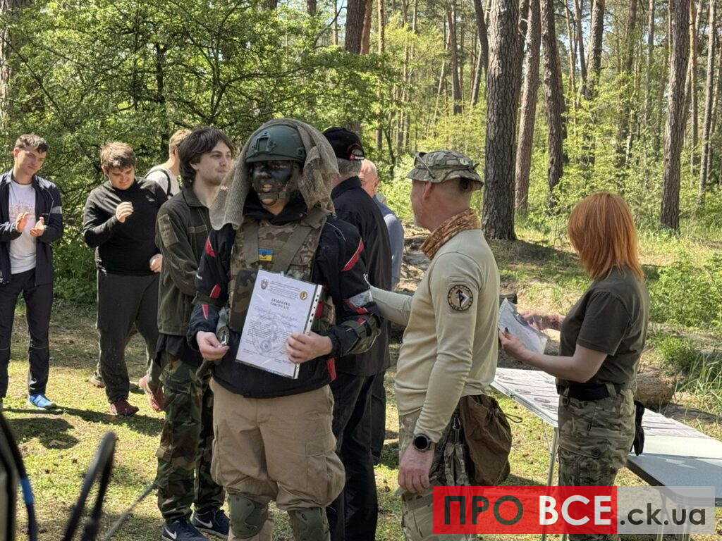 Group of people outdoors in a forest; a person in tactical gear and helmet holds a certificate while a presenter in camouflage speaks nearby.