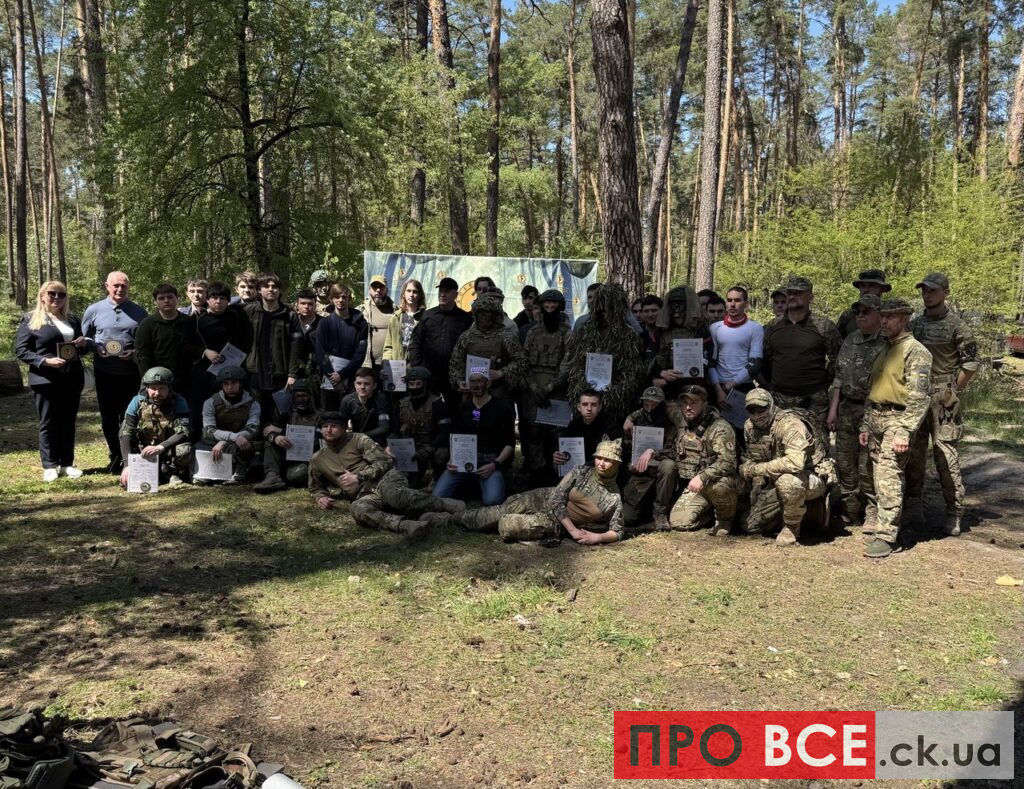 Group of soldiers and civilians posing with certificates in a sunlit forest clearing.
