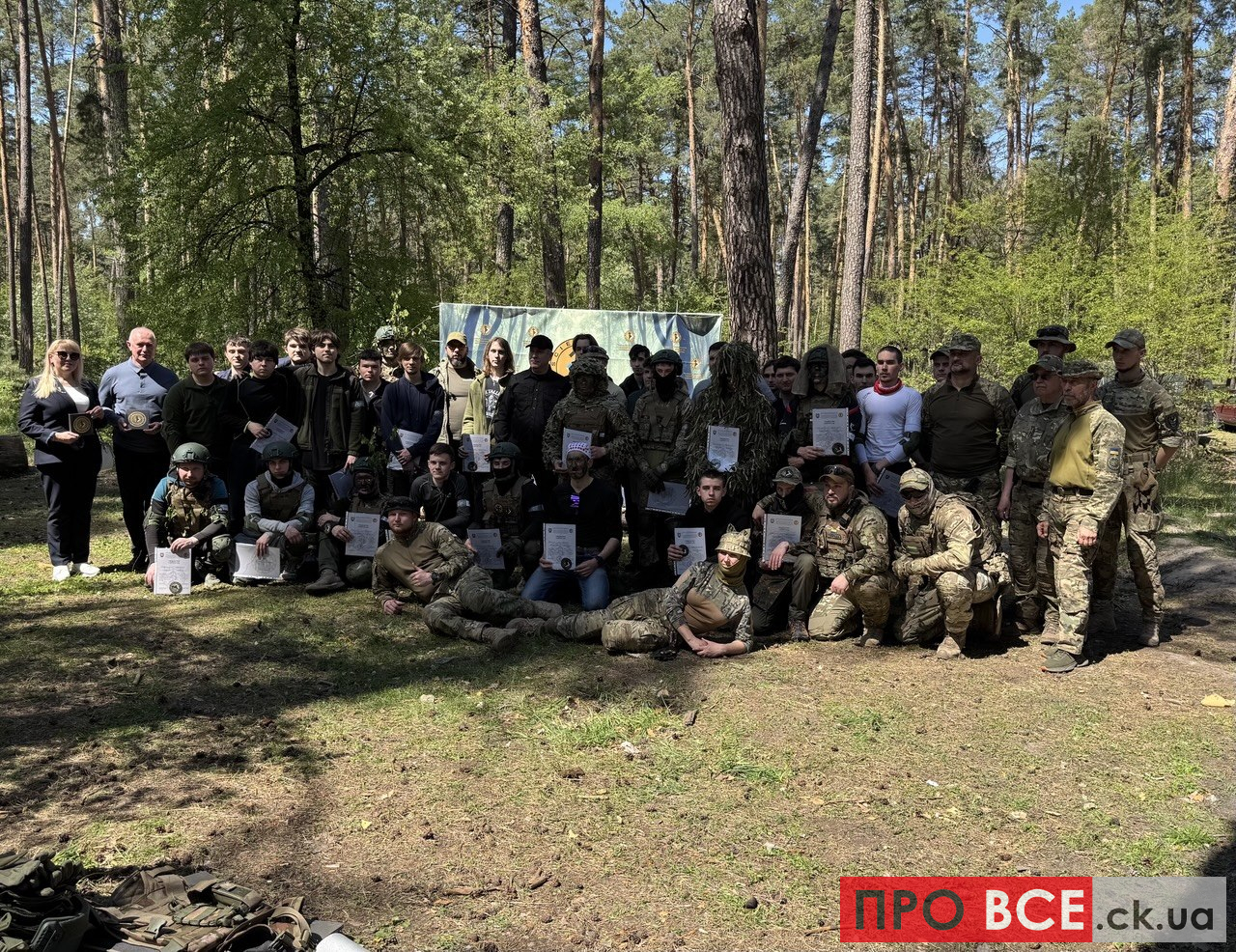 Group of soldiers and civilians posing with certificates in a sunlit forest clearing.