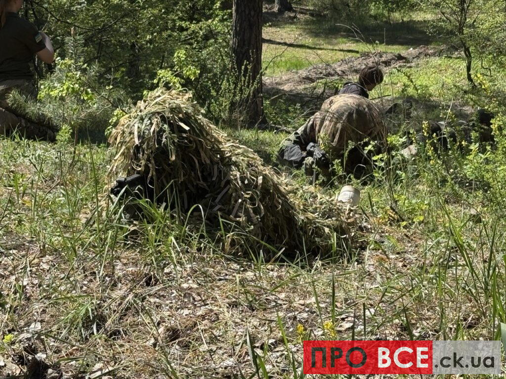 Soldiers in camouflage crouch and take cover in a sunny forest, with a camo net pile and scattered gear nearby.