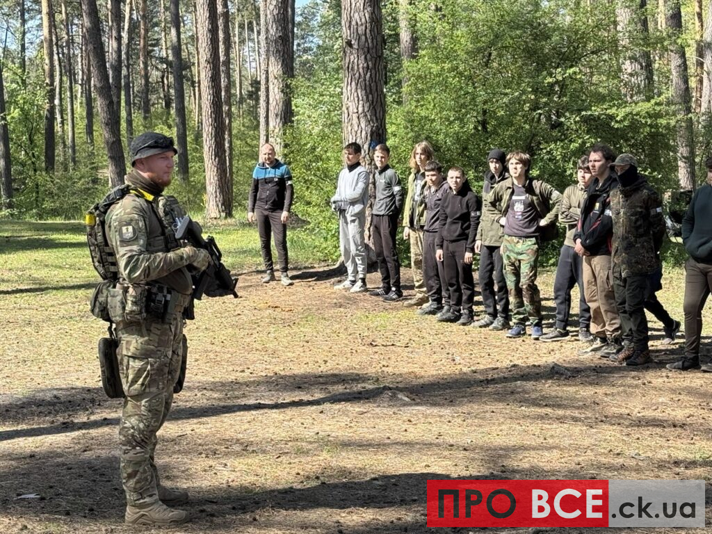 Uniformed soldier with rifle briefing a line of civilians in a pine forest clearing in outdoor training context.
