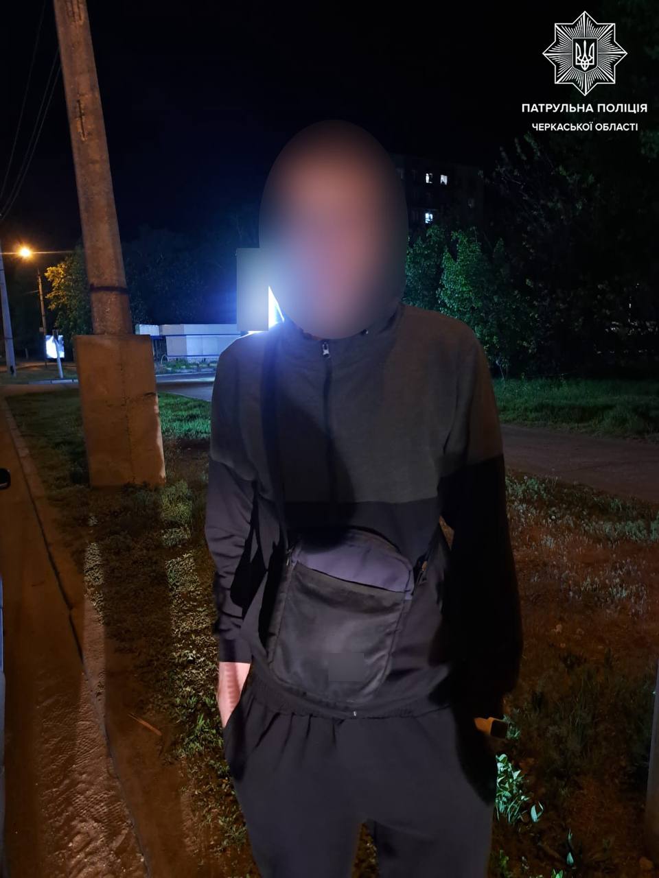 Nighttime street scene with a person wearing a dark hoodie and crossbody bag, face blurred, standing near a curb; police emblem in top-right corner.