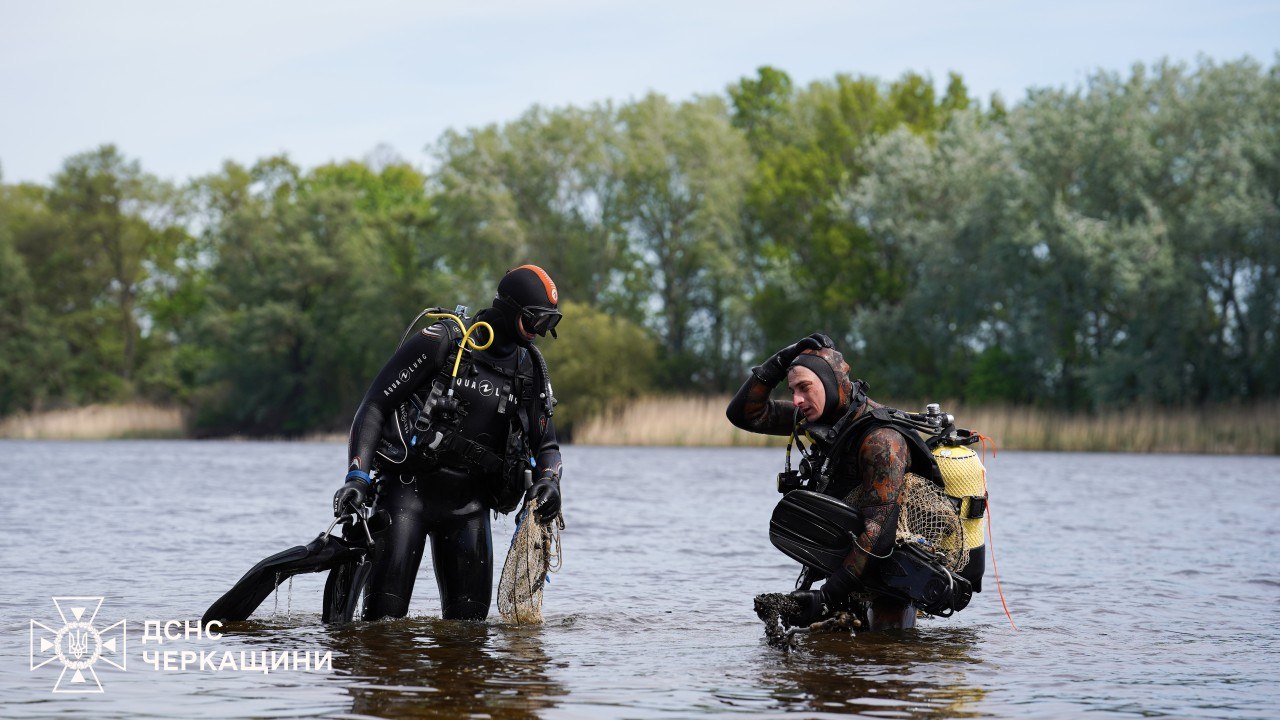 Two divers in wetsuits wading in shallow water, gear on, preparing equipment near a riverbank.