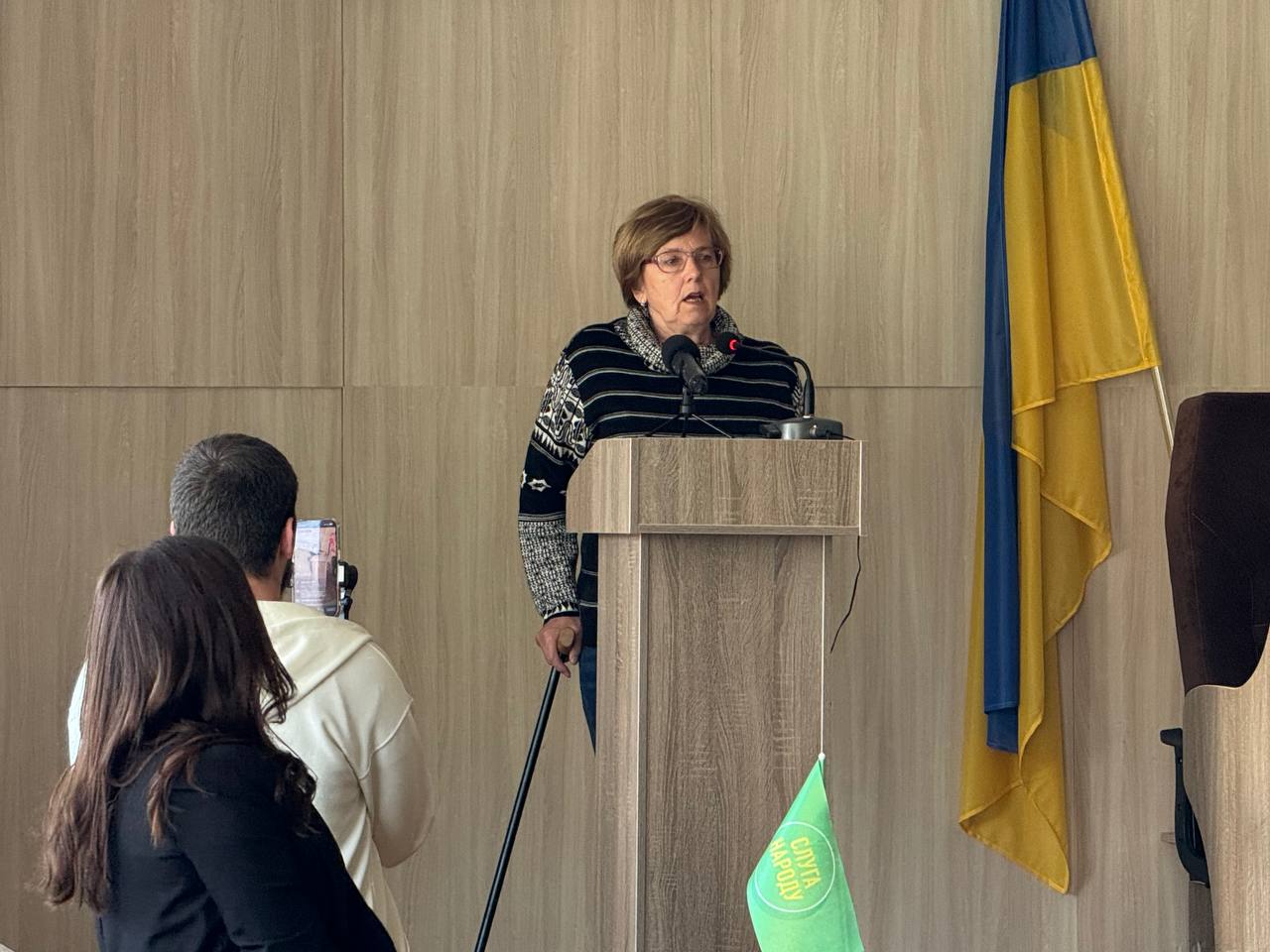 Older woman speaking at a wooden podium with microphones, Ukrainian flag in the background.