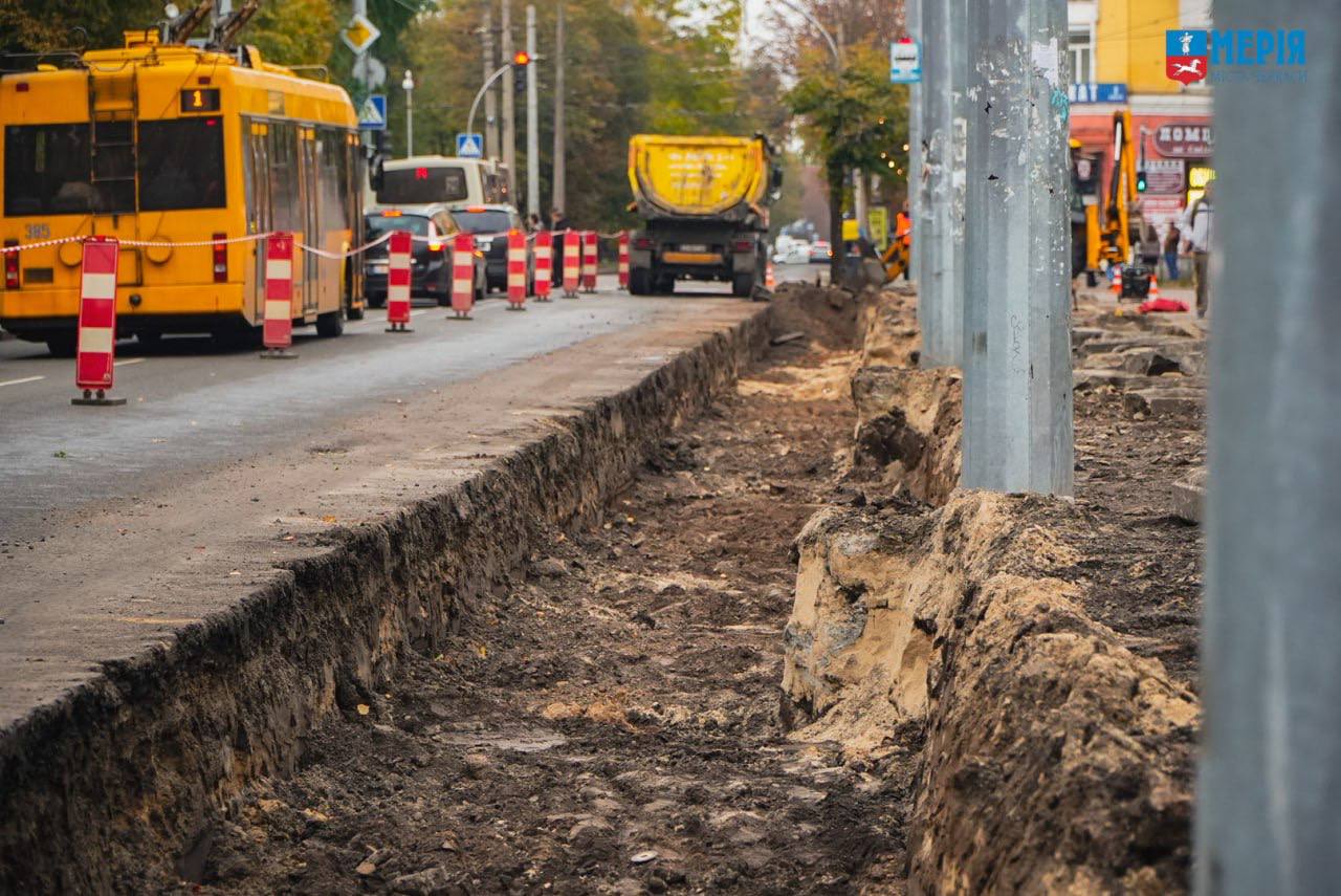 City street under construction with a deep trench along the curb, orange traffic barriers, and yellow buses in the background.
