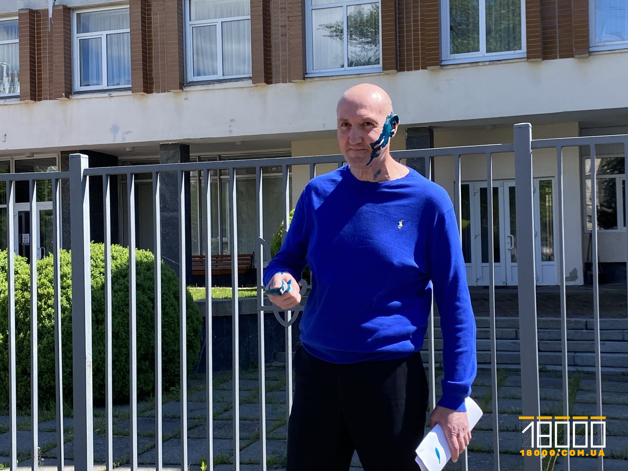Man with blue face and neck paint, wearing a bright blue long-sleeve shirt, standing beside a metal fence in front of a building, holding papers.